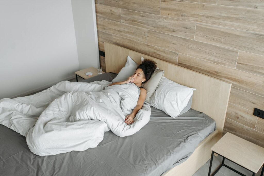A woman lying in bed under a blanket, appearing unwell, in a modern bedroom setting.